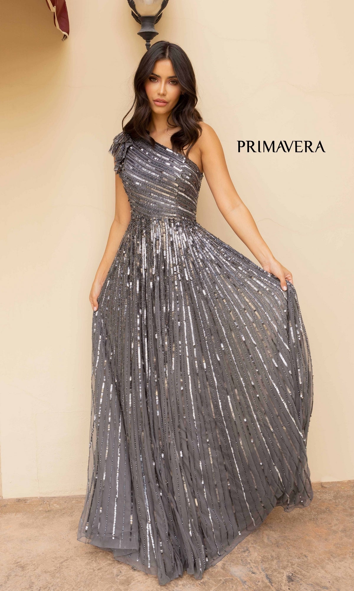 A woman poses in the Strappy-Back One-Shoulder Long Sequin Formal Dress, a floor-length gown with silver sequins and pleats, standing beside a beige wall featuring